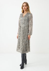 Vrouw in een mouwloze midi-jurk met luipaardprint, gecombineerd met zwarte kniehoge haklaarzen, staand tegen een effen witte achtergrond.