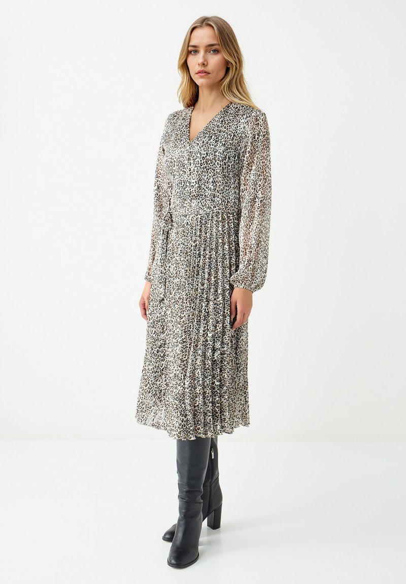 Vrouw in een mouwloze midi-jurk met luipaardprint, gecombineerd met zwarte kniehoge haklaarzen, staand tegen een effen witte achtergrond.