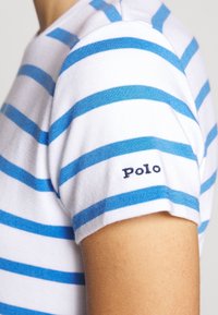 Braccio che indossa una maglietta bianca con righe orizzontali blu e un piccolo etichetta "Polo" sulla manica.