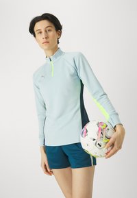 Top sportivo di colore azzurro chiaro con colletto alto e dettagli verde neon, abbinato a pantaloni corti blu scuro. Una palla da calcio con motivi colorati è tenuta in mano.