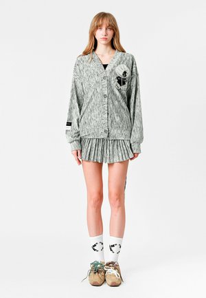 Cárdigan de punto gris con cinco botones, tejido texturizado, falda plisada y calcetines con estampado. Con detalle de flor y rayas.