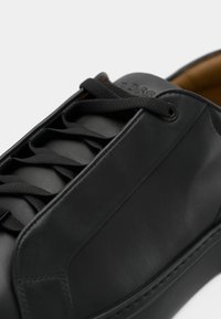 Black leather sneaker with black laces, visible stitching, and padded brown interior lining.