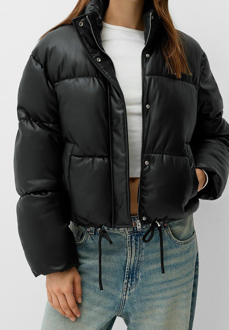 Svart kort puffjacka med glänsande yta, tryckknappsstängning och dragsko i nederkant. Matchad med ljusblå denimjeans.