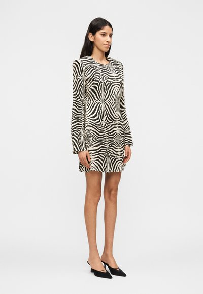 Γυναίκα με φόρεμα ζέβρας μέχρι το γόνατο με μακριά μανίκια, φορώντας μαύρες μυτερές γόβες, στέκεται μπροστά σε λευκό απλό φόντο.