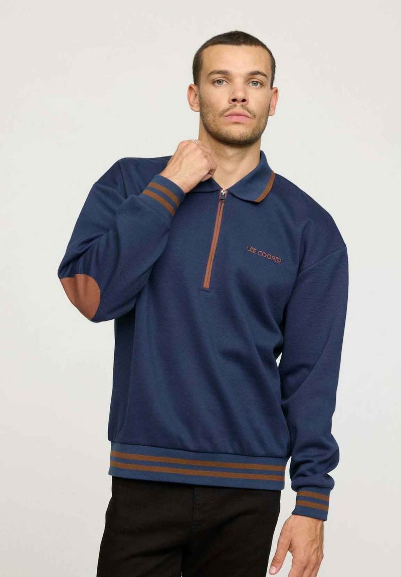 Pull-over bleu marine avec un col zippé demi, des rayures marron et des coudières. Présente une coupe décontractée et un logo brodé sur la poitrine.