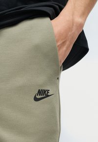 Άτομο που φοράει χακί πράσινο φούτερ Nike με το χέρι στην πλαϊνή τσέπη και μαύρο τοπ μερικώς ορατό.