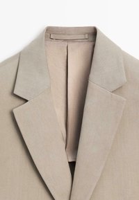 Beige colbertkraag en revers met zichtbare stiksels en een stoffen lus binnenin de halslijn tegen een witte achtergrond.