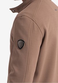 Brun struktureret jakke med et ribbet design, der har en høj krave og et sort logo-patch på venstre skulder.