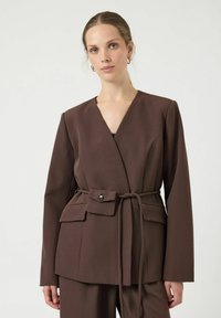 Bruine wikkelblazer met V-hals, lange mouwen en een ceintuur die gestrikt kan worden. Heeft een klepzak en een gladde stoftextuur.