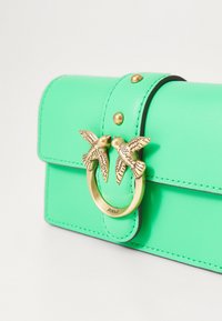 Geantă de piele verde cu închidere cu clapetă, având un inel din metal auriu și două accente sub formă de păsări. Suprafețe texturate cu cusături contrastante.