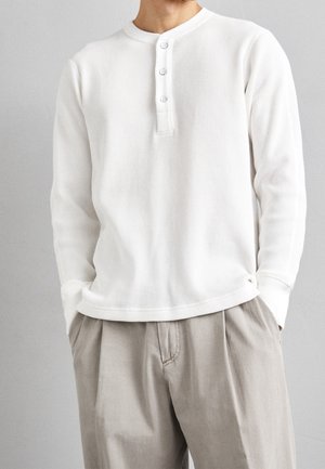 Homme portant une chemise henley blanche à manches longues en tricot gaufré avec trois boutons et un pantalon beige ample, mains dans les poches, fond neutre.