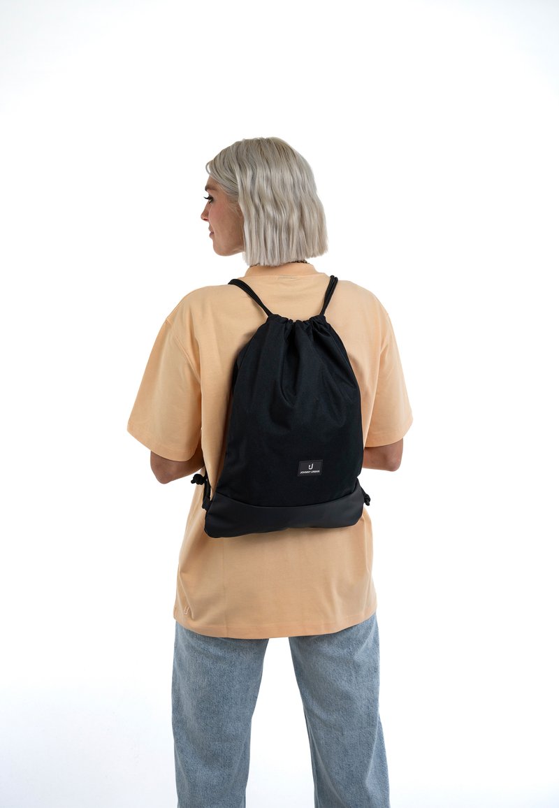 Persona con capelli biondi corti che indossa una camicia beige e jeans azzurri, con uno zaino nero con coulisse, vista da dietro.