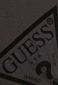 Tmavě šedé bavlněné tričko s velkým černým trojúhelníkovým logem, na kterém je nápis "GUESS" a "U.S.A.", spolu s designem otazníku.