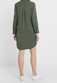 Šaty s dlouhým rukávem v olivově zelené barvě, s límečkem, zadním skladem a bočními rozparky, doplněné o bílé tenisky. Jemná textura látky.