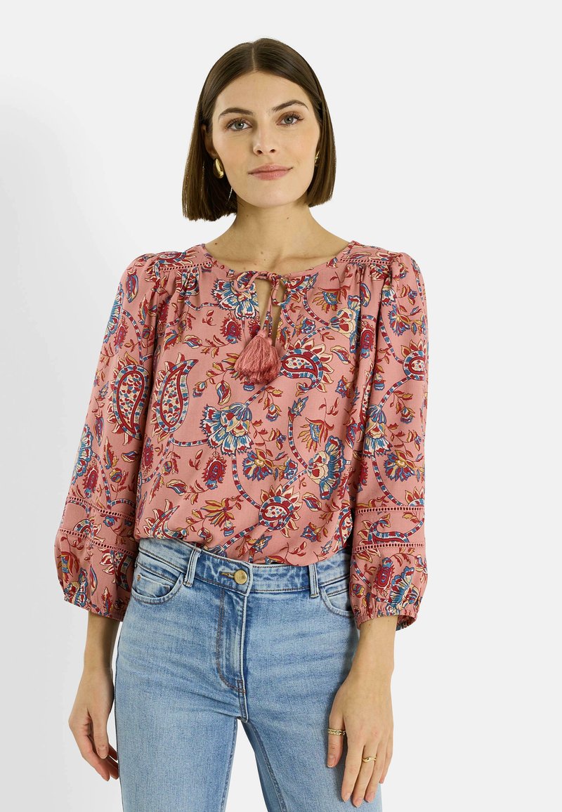 Mujer con cabello castaño lacio, lleva pendientes de aro dorados, una blusa rosa con estampado floral y tasseles, y jeans azul claro, de pie con las manos relajadas.