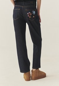 Sötétkék farmernadrág egyenes szabásban, hátul hímzett virág- és madármintákkal a zsebeken. Barna bokacsizmával párosítva.