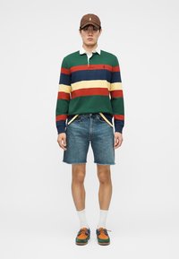 Άνδρας που φορά καφέ καπέλο, μακρυμάνικη ράγκμπι μπλούζα με πράσινες, κόκκινες, ναυτικές και κίτρινες ρίγες, τζιν σορτς, λευκές κάλτσες και πολύχρωμα παπούτσια.