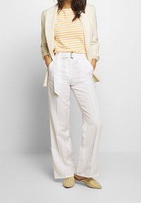White wide-leg trousers with a belted waist, paired with a yellow and white striped top underneath a light beige blazer. Woven slip-on shoes.