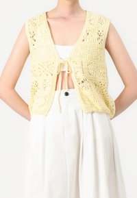 Keltainen neulottu liivi avoimella edessä, monimutkaisilla pitsikuviolla, lyhyillä hihoilla ja edessä solmittavalla yksityiskohdalla, puettuna valkoisen topin ja hameen päälle.