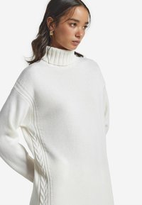 Weißer Strickpullover mit hohem Rippkragen, seitlichem Zopfmuster und lockerer Passform. Weiche Textur mit langen Ärmeln.