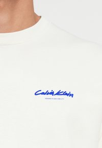 Sweatshirt blanc en coton, arborant un logo brodé bleu sur la poitrine gauche avec le texte « Calvin Klein » et « CONÇU À NEW YORK CITY ». 