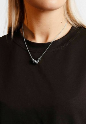 Silverkedja med tre dekorativa metallpärlor på en svart t-shirt med rund halsringning, som visar upp en elegant och minimalistisk design.