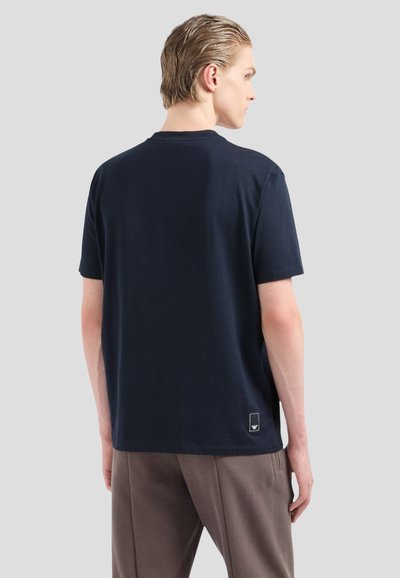 T-shirt en coton bleu marine à manches courtes, col rond, avec une petite étiquette de marque en bas à gauche au dos. Texture lisse.