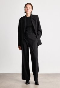 Melna uzvalka bikses ar piegrieztu žaketi ar vienu pogu, teksturētu melnu džemperi un plati bikses. Gluda auduma apdare.