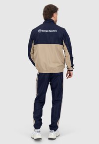 Tuta navy e beige con giacca con zip e polsini elastici. Presenta il logo sulla parte posteriore e strisce laterali sui pantaloni.