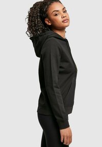 Svart zip-up hettegenser laget av mykt stoff, med kenguru-lomme, ribbestrikkede mansjetter og hette med snor. Slank tekstur og tettsittende design.