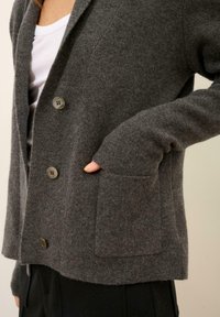 Szary wełniany kardigan z wyciętym kołnierzem, trzema guzikami i przednią kieszenią z prążkowanym rękawem. Miękka tekstura i luźny krój.