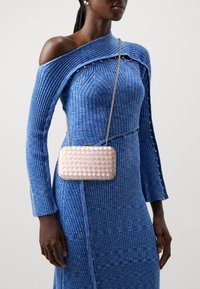 Różowa torebka z plecionego materiału z metalowym zapięciem i łańcuszkiem na ramię, trzymana na wysokości talii, zestawiona z prążkowaną niebieską sukienką z szerokim kołnierzem.