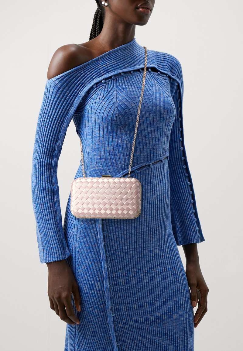Różowa torebka z plecionego materiału z metalowym zapięciem i łańcuszkiem na ramię, trzymana na wysokości talii, zestawiona z prążkowaną niebieską sukienką z szerokim kołnierzem.