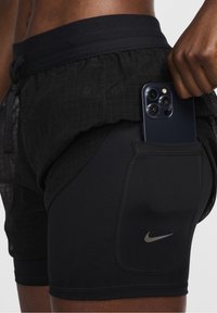 Pantalones cortos deportivos negros con una capa exterior de malla, cinturilla elástica y un forro interno ajustado; cuenta con un bolsillo lateral que sostiene un teléfono inteligente.