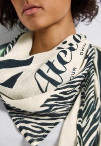 Närbild av en person som bär en krämf�ärgad och svart mönstrad scarf med fet text och zebraränder, lindad runt halsen.