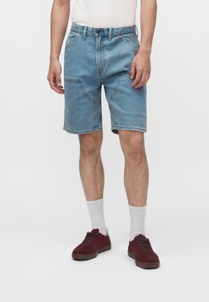 Homme portant un short en jean bleu clair avec de grandes poches devant, des chaussettes blanches et des baskets rouge foncé, debout sur un fond blanc.