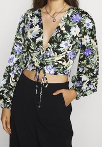 Top corto floreale verde, con fiori viola e blu, scollatura a V profonda, maniche lunghe a sbuffo e un laccio in vita. Pantaloni neri indossati sotto.