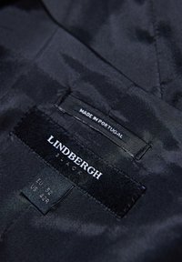 Czarna etykieta z tkaniny z napisem "LINDBERGH BLACK" w białym kolorze, z oznaczeniami rozmiarów EU 52 i US 42R, z informacją "WYPRODUKOWANO W PORTUGALII."