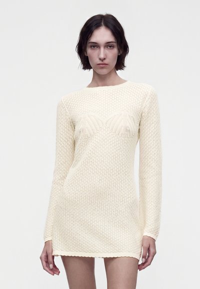 Femme aux cheveux courts et foncés portant une robe en tricot beige crème, ajustée, à manches longues, se tenant devant un fond blanc uni.