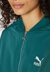 Teal bomberjakke med lynlås foran, ribbet krave og satinfinish; har et lille hvidt Puma-logo på venstre bryst.