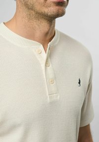 Hvit teksturert kortermet Henley-skjorte med tre knapper og en liten marineblå logo på venstre bryst. Ribbet krage og mansjetter.