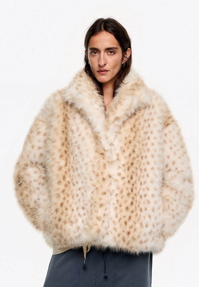 Chaqueta de piel sintética beige con una textura suave, que presenta un amplio collar, un patrón de lunares y un corte holgado. Detalle de cordón en el dobladillo.