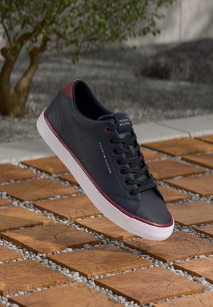 Baskets synthétiques bleu marine avec une texture lisse, accents rouges, semelle blanche et lacets noirs. Présente un logo sur le côté.