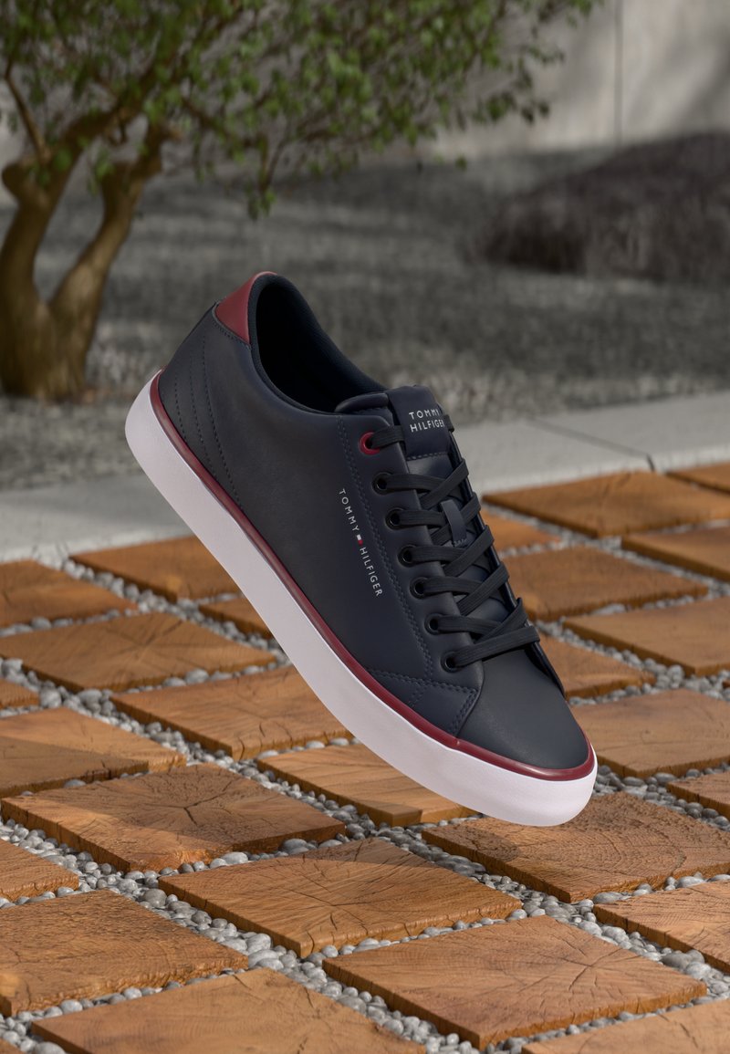 Zapatilla sintética de color azul marino con textura suave, detalles en rojo, suela blanca y cordones negros. Presenta una marca en el costado.