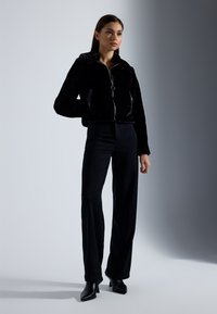 Zwarte nepbont jas met een ritssluiting aan de voorkant, grote kraag en zijzakken, gecombineerd met zwarte slim fit broek en puntige haklaarzen.