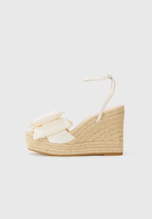 Wedge sandal with a natural jute platform, cream fabric upper, and two layered floral accents; features a thin ankle strap with buckle.