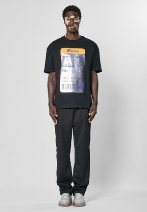 Svart grafisk T-skjorte med design av en flybillett, kombinert med svarte joggebukser og grå sneakers med en gjennomsiktig såle.