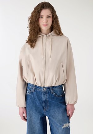 Giovane donna con capelli ricci che indossa una giacca beige con cappuccio e cerniera e jeans blu a vita alta con ginocchio strappato.