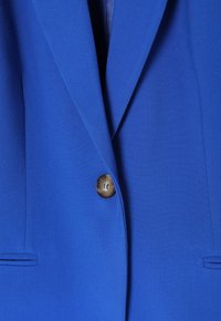 Modrý blejzr s jedním knoflíkem, ostrými klopami a bočními kapsami z hladké texturované látky. Knoflík má kontrastní design.