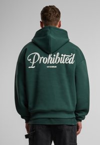 Sudadera con capucha de tejido verde oscuro. Presenta un gran texto blanco "Prohibido" en la parte posterior, con "10119 Berlín" debajo en tamaño más pequeño. Puños y dobladillo acanalados.